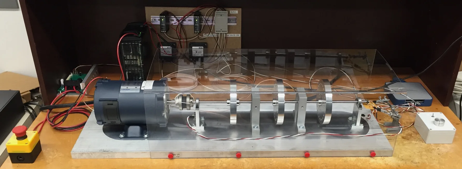 Rotor imbalance test rig: a motor coupled through a flexible coupling to an instrumented shaft carried in four bearing blocks on an aluminium base plate, with data acquisition modules on the panel behind and an emergency stop at the near end.
